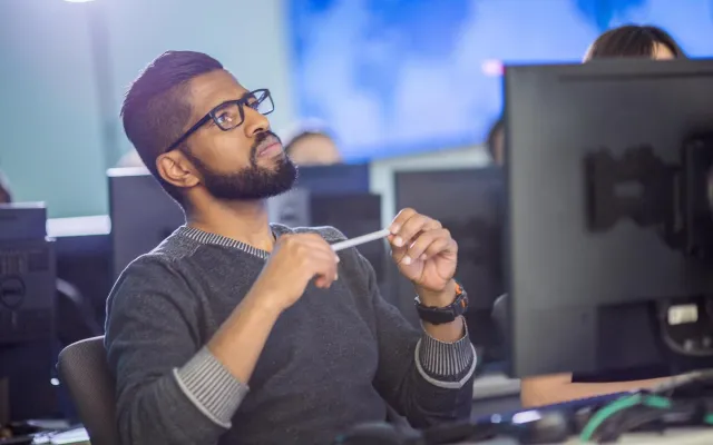 Student with beard and glasses sitting at a computer monitor holding a pen