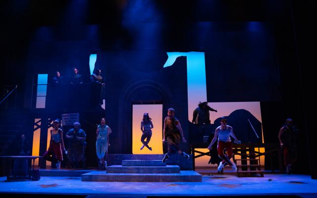 A scene from the University of Lethbridge's production of Jesus Christ Superstar. A number of actors hovering in the air mid-dance. Set is cast in a darker blue light. Backdrop is backlit with lighter blue, magenta and yellow.