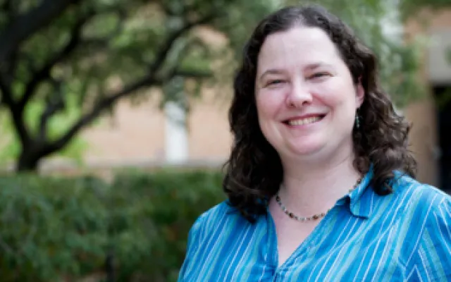 A white woman with wavy shoulder-length brown hair, freckles, and a blue and white shirt is smiling outside of University Hall. An oak tree and a hedge are in the background