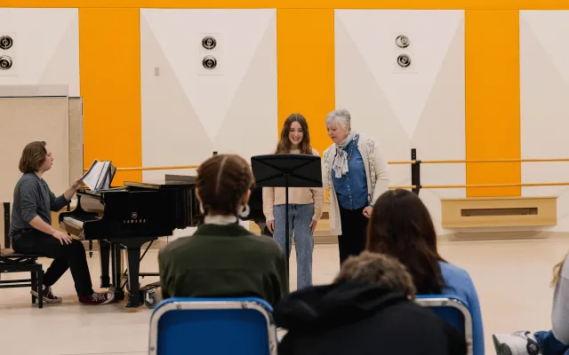 a voice professor working with a student in a music workshop with an accompanist sitting at a grand piano.