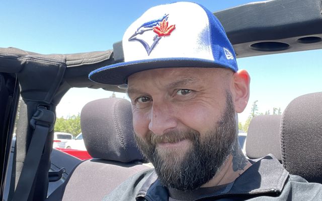 Picture of a caucasian man with mustache and beard, wearing a blue and white ball cap while sitting in an open-topped Jeep.