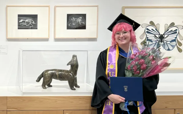 Picture of Jessica Colley, a young woman with pink hair, in a graduation cap and robe, holding flowes and a degree in a blue folder, standing next to an art gallery display of framed pictures and a sculpture.