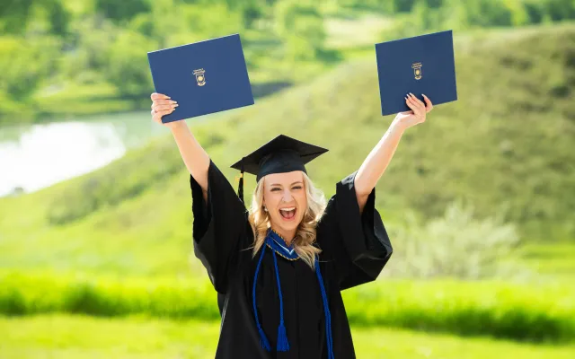 Graduate in front of a green background, holding up two degrees in celebration