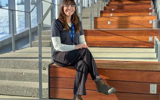 Brunette woman sitting on a bench next to stairs. Her left leg is crossed over her right. She is wearing glasses, a grey shirt, and a long sleeve undershirt. She wears a blue Agility lanyard.