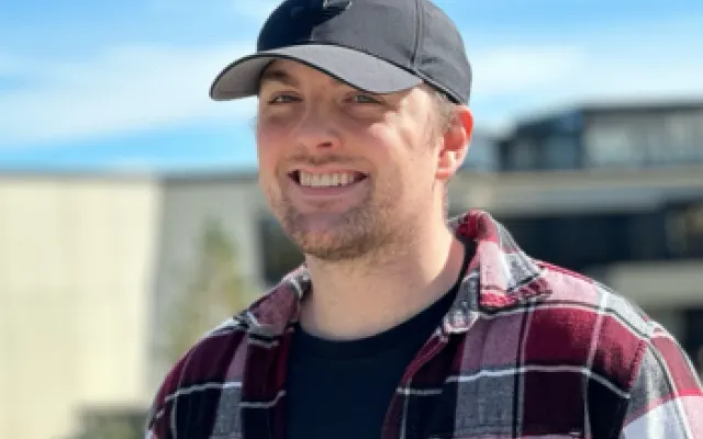 Photo of Clay with a black baseball cap on in front of the front entrance to the library. It's a sunny day and he is smiling.