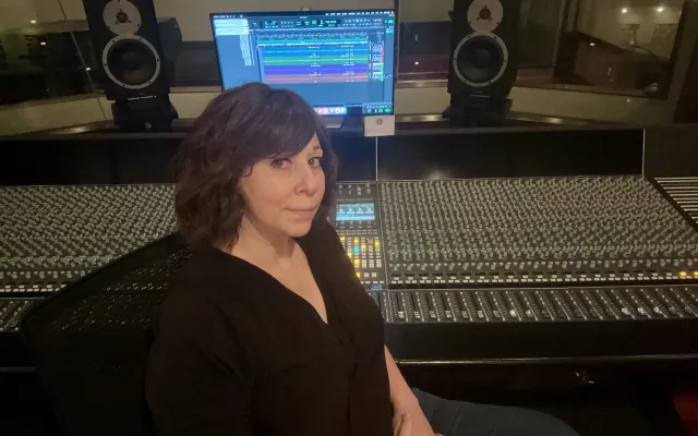 A photograph of Mary Mazurek from the waist up, sitting before a large control panel in a recording studio.  She is a Caucasian woman with dark, shoulder-length hair.