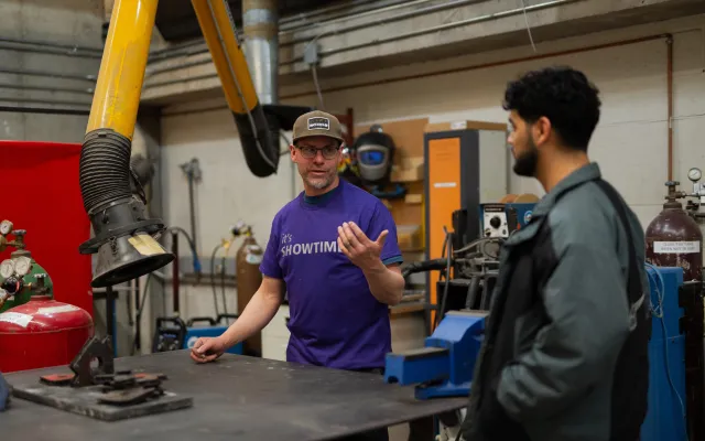 Volunteer in purple tshirt talking to another man in a construction workshop.