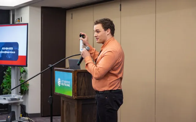 A man holding a prop bear spray canister while holding a microphone, standing in front of a podium