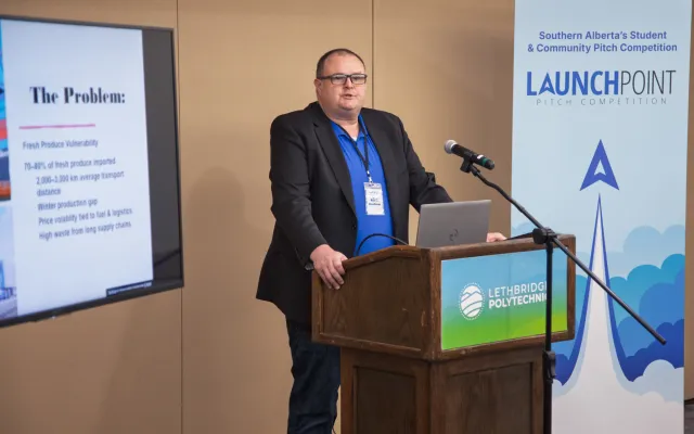 A man wearing a jacket stands behind a mic and podium, in front of a screen with a presentation and a banner.