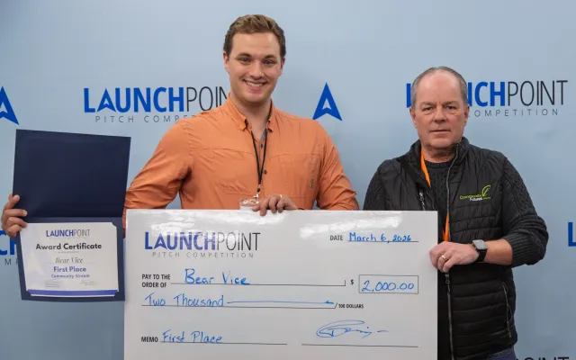 Young man standing next to an older man in front of a blue backdrop, holding a big cheque addressed to Bear Vice and a certificate.