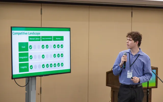 A young man holding a microphone, standing in front of and looking towards a presentation slide on a tv screen