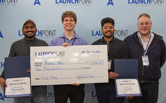 Four men standing in front of a blue backdrop. One holds a big cheque, the two on either side hold small certificates. They are all smiling.