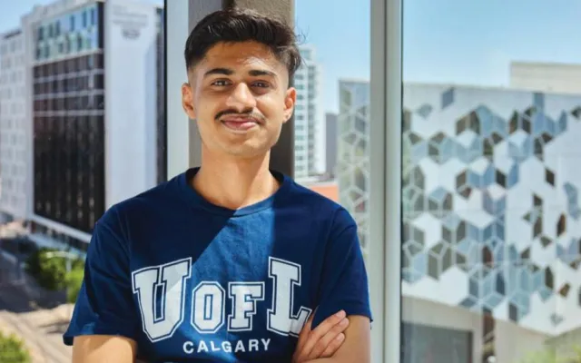 A photo of Habil wearing a U of L Calgary T-Shirt infront of a window showing Downtown Calgary from the ULethbridge Calgary Campus