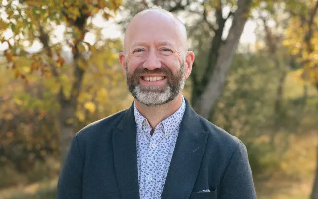 A man in a suit, standing in front of a group of trees