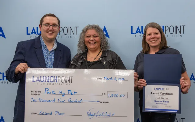 Three people standing in front of a blue background, two staff one student, holding a large cheque and a certificate