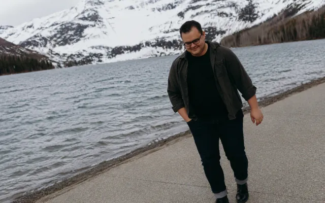 Colour photograph of Jude Bialik - a man in dark clothing with short hair and glasses - walking alongside a body of water, with a snowy mountain in the background.