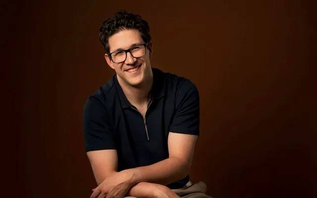 Picture of Alex Bohn, a caucasian man with short, dark hair and glasses, wearing a dark shirt, seated with forearms crossed in front of a brown background.