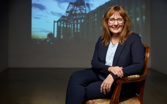 A photograph of Anne Dymond - a smiling woman with long, light brown hair, glasses, and a dark blue suit.  She is seated on an upholstered wooden chair, in a dark room with a slightly blurry architectural image projected on the wall behind her.