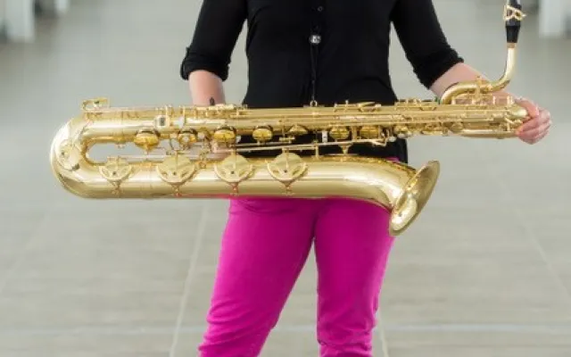 A picture of Holly De Caigny - a cauciasian woman with mid-length brown hair, wearing a black shirt and pink pants, holding a baritone saxophone, standing in a long hallway.