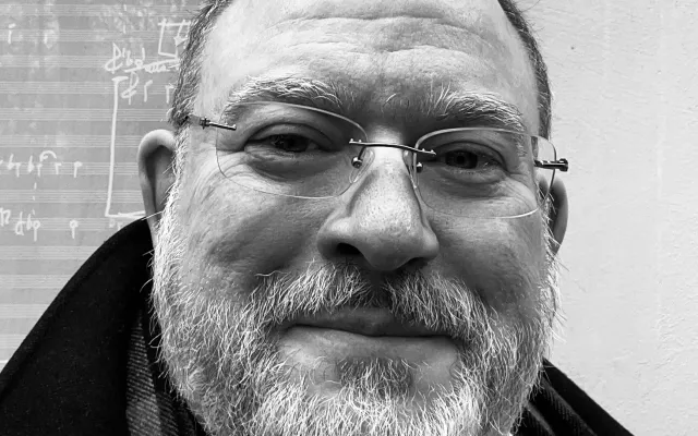 A black and white photograph of Dr. Arlan Schultz from the neck up, in front of a reflective window.  He is a caucasian man with short hair, a mustache, and beard, and is wearing rimless eyeglasses and a dark jacket.