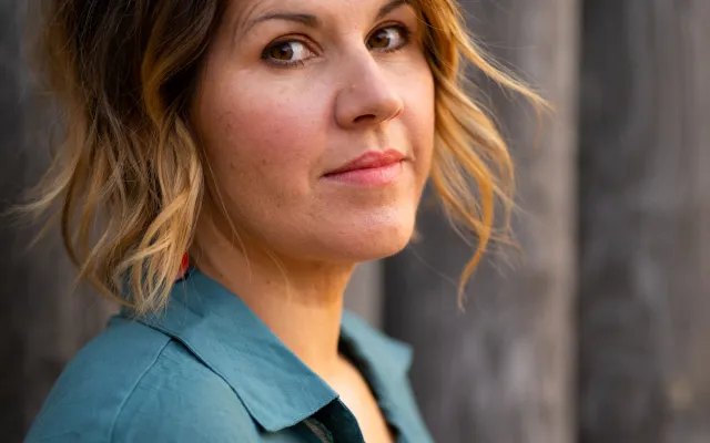 A photograph of Nicola Elson - a caucasian woman with mid-length hair, dark at the roots and lighter brown at the ends, pictured from the chest up in 3/4 profile.  She is wearting a blue blouse, and is standing in front of an unfocused, pillar-like bakground.