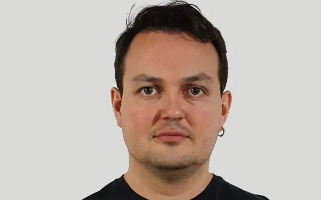 man with an earring in a black shirt in front of a plain wall