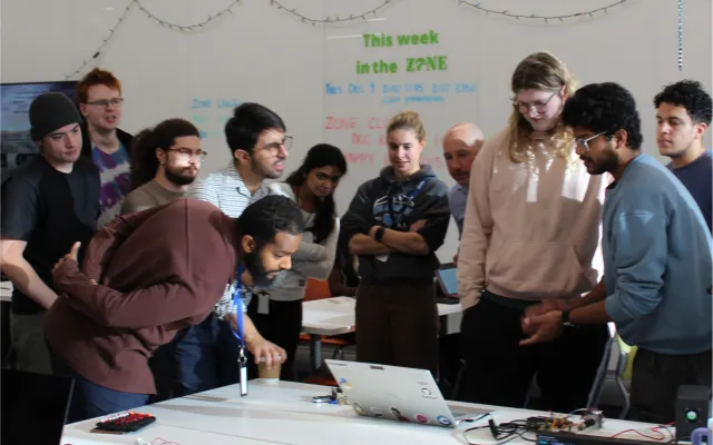 a group of students and their instructor huddled around a piece of electronics equipment and a laptop. One student is explaining the process.