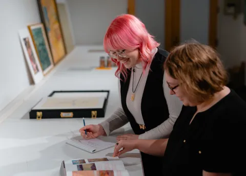 Jessica Colley (left) and Kate Riddoch (right). Two University of Lethbridge museum studies intern students smiling while looking through an art textbook and writing notes.