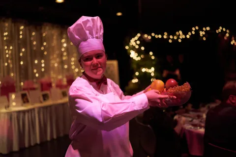 A person in a chef hat and apron carrying a bowl of fruit.