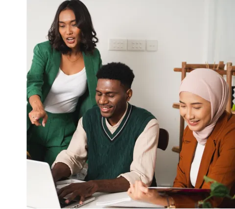 Three young adults sitting around a laptop, all smiling, while one is points over the shoulder of another at the laptop screen