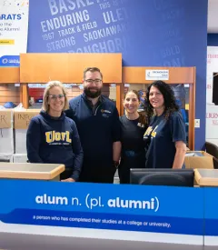 Four ULethbridge staff stand behind a sign that says, "alum n. (pl. alumni)" and smile.