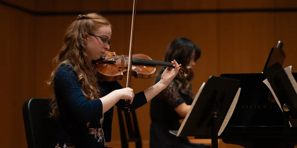Alayna McNeil is seated playing the violin and Anna Jeong is seated playing the black grand piano.