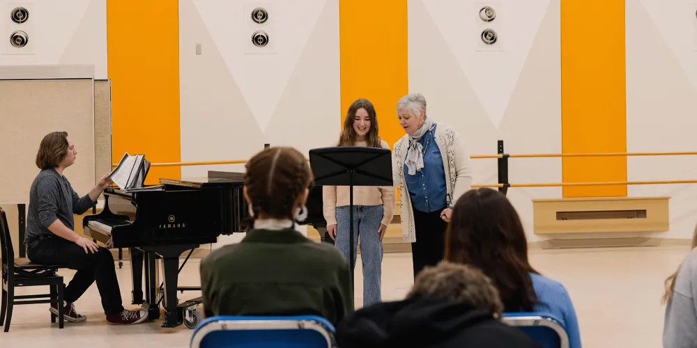 a voice professor working with a student in a music workshop with an accompanist sitting at a grand piano.