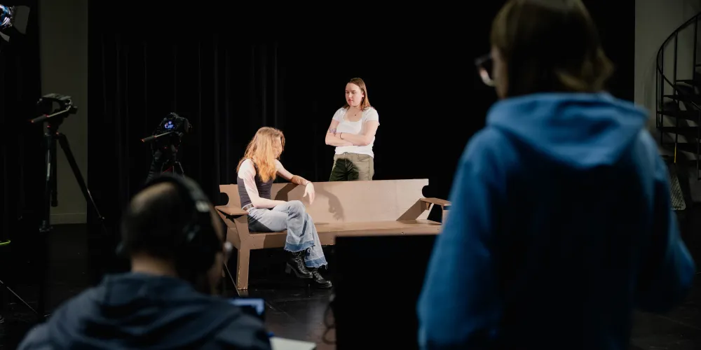 Students working in front of and behind the scenes in an acting for the camera class. Highlighted is a student is sitting on a bench and another is standing behind the bench with arms crossed while looking at the seated person. The camera crew and equipment are blurred slightly in the foreground.