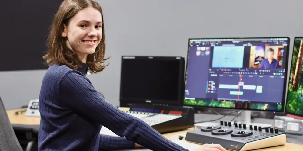 A woman in a dark blue long sleeve top sits in front of a computer with video editing equipment.