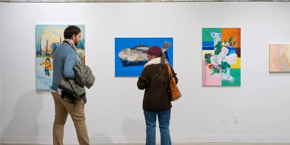 A masculine person and a femme person in an art gallery looking at a poster-sized beautifully painted fish on a vibrant blue background