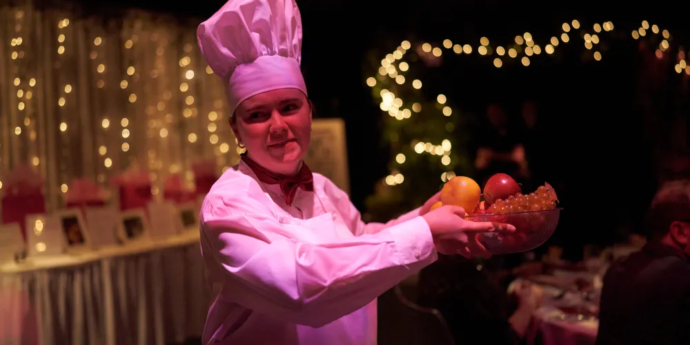 A person in a chef hat and apron carrying a bowl of fruit.