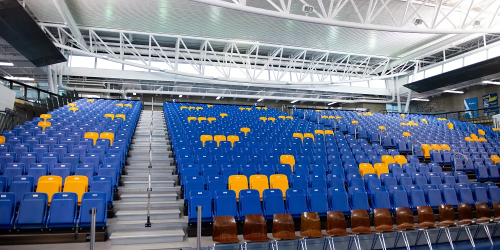 A wide-lens photo of empty bleacher seating. Most of the chairs are blue, but there are yellow ones sporadically throughout. All of the chairs fold down except for a row of chairs with legs on the floor of the gym.