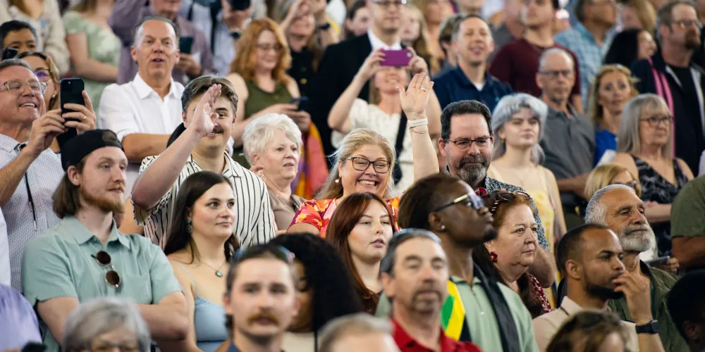 A side-angle photograph of dozens of guests sitting in the bleachers as they watch the ceremony. Some hold up their phones as they record.