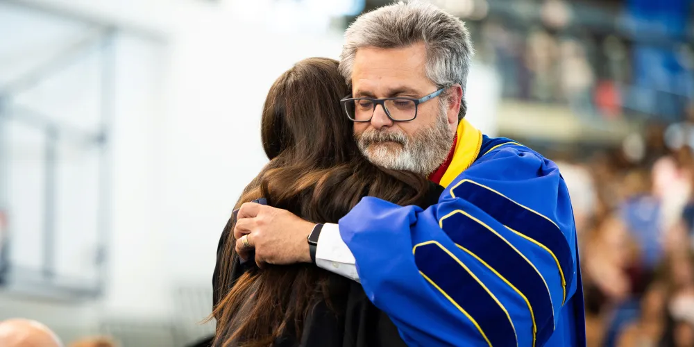 A grad hugs a faculty member as they cross the stage. The grad's back is to the camera. The faculty member wears blue regalia with dark blue striped patches on the arms.