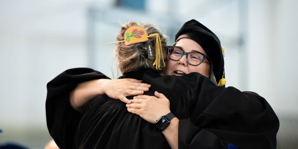 A grad hugging a faculty member as they cross the stage. The grad's back is toward the camera.