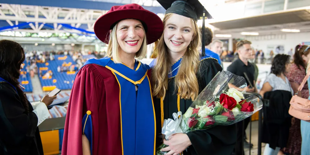 A grad stands next to a faculty member, who is dressed in red, blue, and gold regalia. The grad is holding a bouquet of flowers.