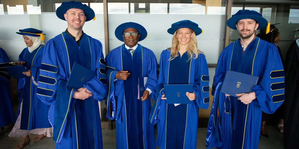 Four PhD grads dressed in blue regalia and holding their degrees