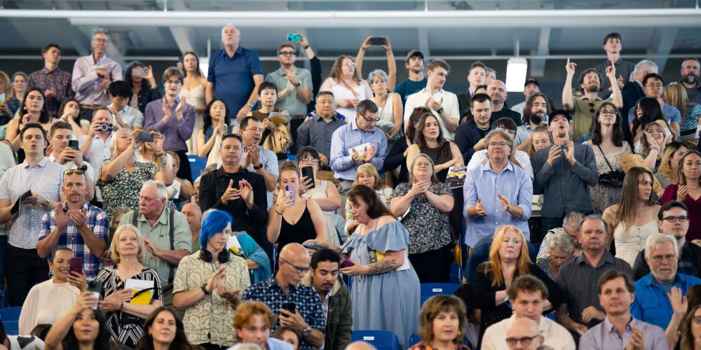 Dozens of guests stand in the bleachers and face the camera