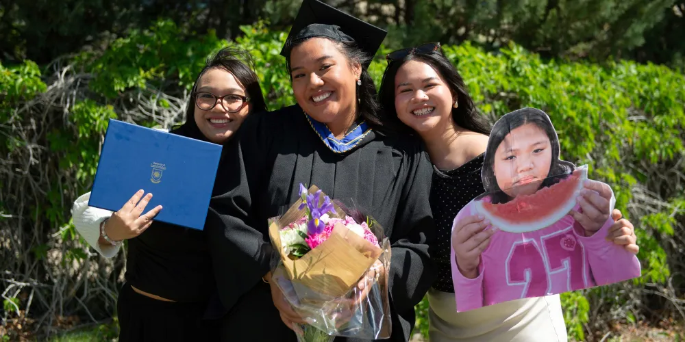 Three women smiling and each holding an item: degree, bouquet, and cut-out image of the grad