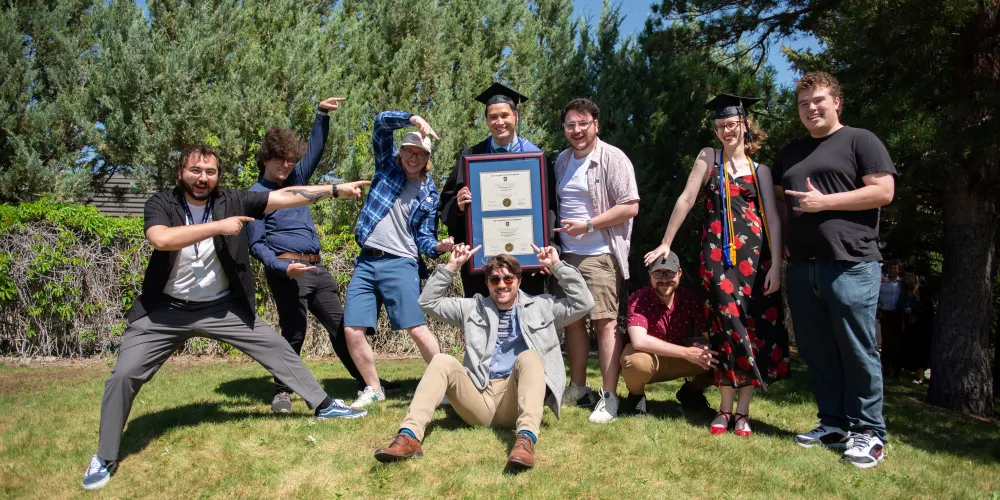 A fun group photo taken on a grass hill. The grad stands in the middle and holds a frame with two parchments. Their guests are pointing toward them and doing fun poses.