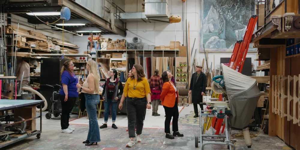 A group of people walk through a theatre scene shop.