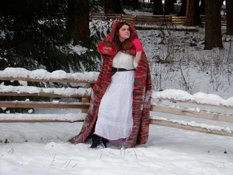 photo of Olympia Tomasta wearing a white dress and red hooded cape standing in a snowy background.
