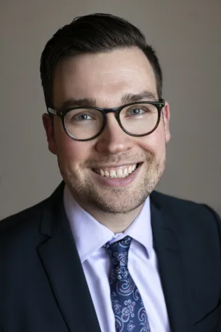 Smiling young man with brown hair and dark frame glasses, wearing a dress shirt and paisley tie.