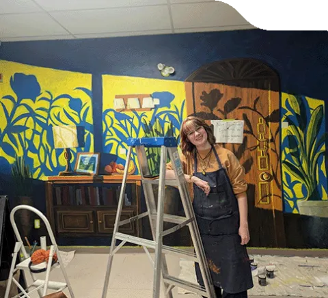 Photo of Kennedy Chinn leaning on a step ladder in front of her mural.